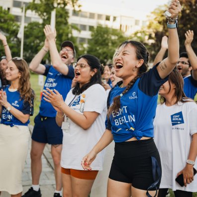 ESMT Berlin Full-time MBA students Ting Han, Roshni Dutta, Tammy Sokari, Jasmin Schubach and Christopher Gerbino cheering during the school Olympics. They are all wearing ESMT-brand tee shirts and are standing in the campus garden on a sunny evening.