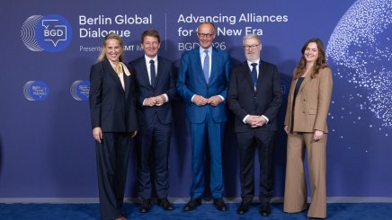 Five attendees pose in suits before Berlin Global Dialogue blue backdrop.