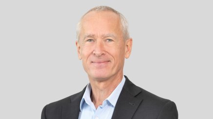Professional headshot of smiling gray-haired man in suit against gray background.