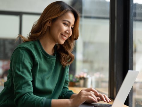 Woman in her early thirties sitting in a cafe and typing on her laptop. She has long brown hair and a silky emerald shirt.