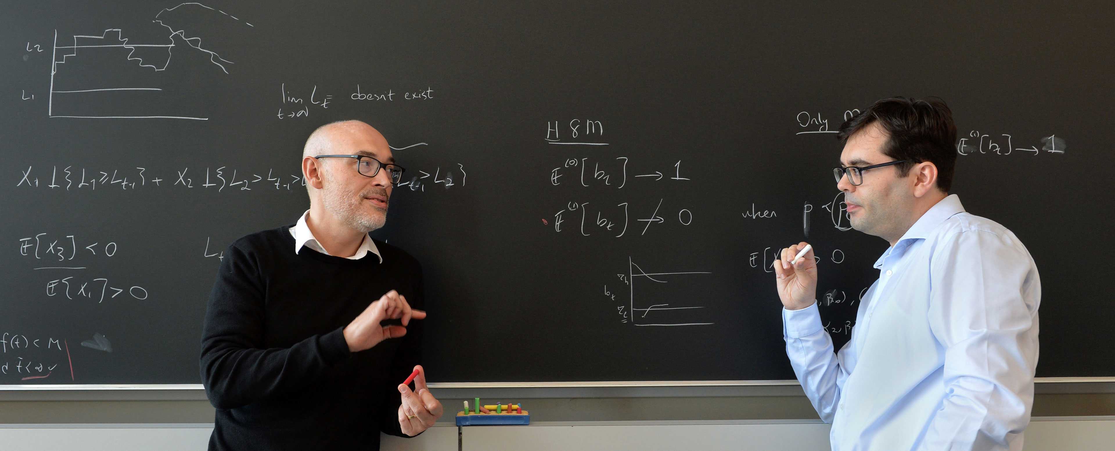 2 male professors standing in front of a blackboard having a discussion