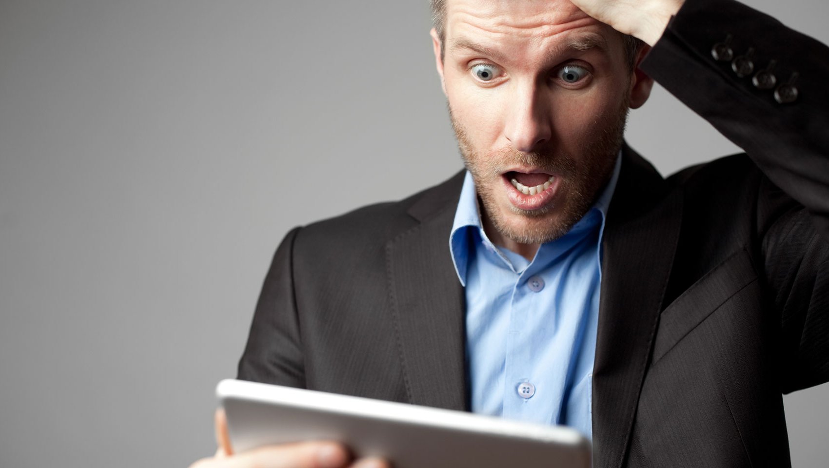 Whie male in a business suit with a shocked expression on face looking at tablet. 